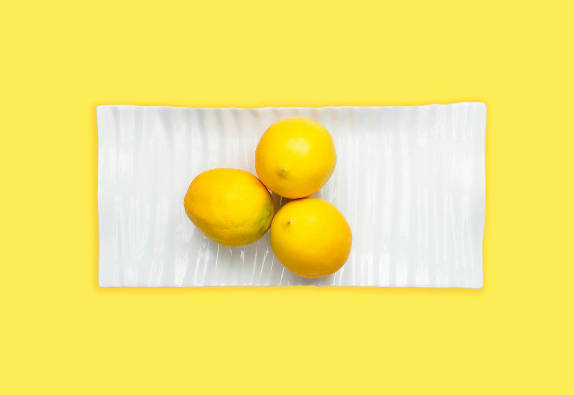 Three fresh lemons arranged on a white plate with a vibrant yellow background