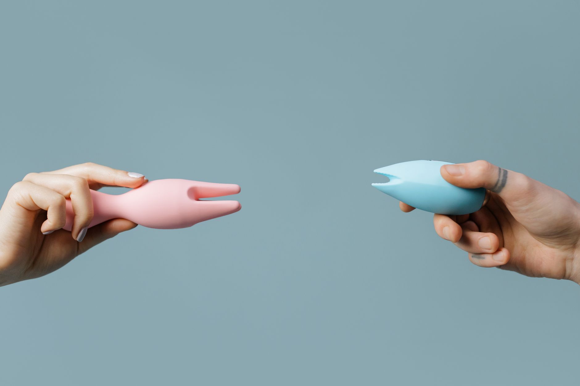 Two hands holding pink and blue silicone clitoral vibrators against a pastel background, representing intimate wellness options for postpartum recovery.