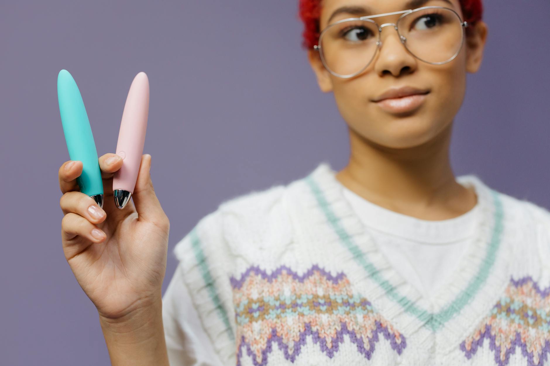 A young woman with eyeglasses holds two colorful vibrators against a purple background