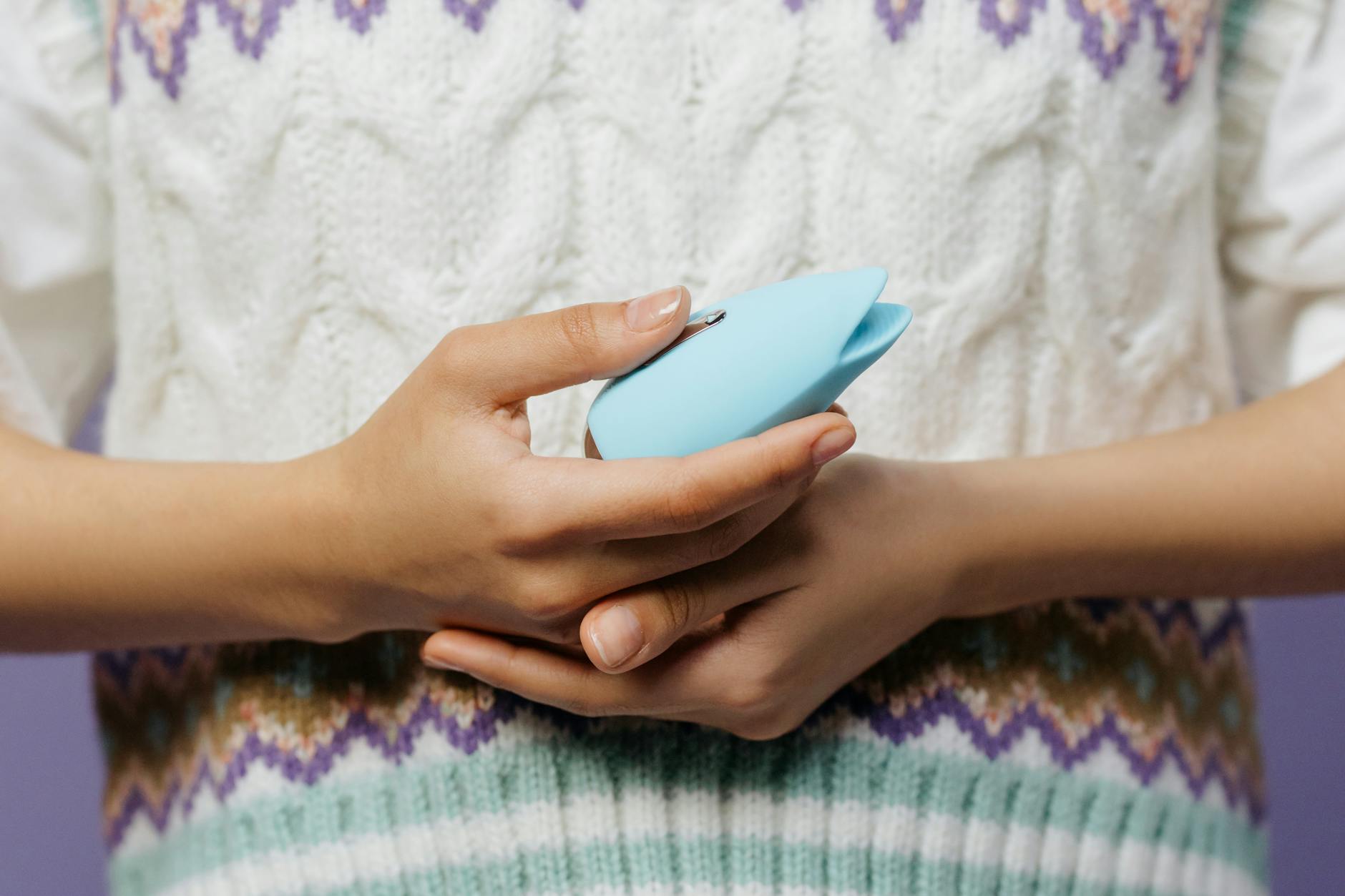 Close-up of hands holding a sleek blue clitoral vibrator against a purple background