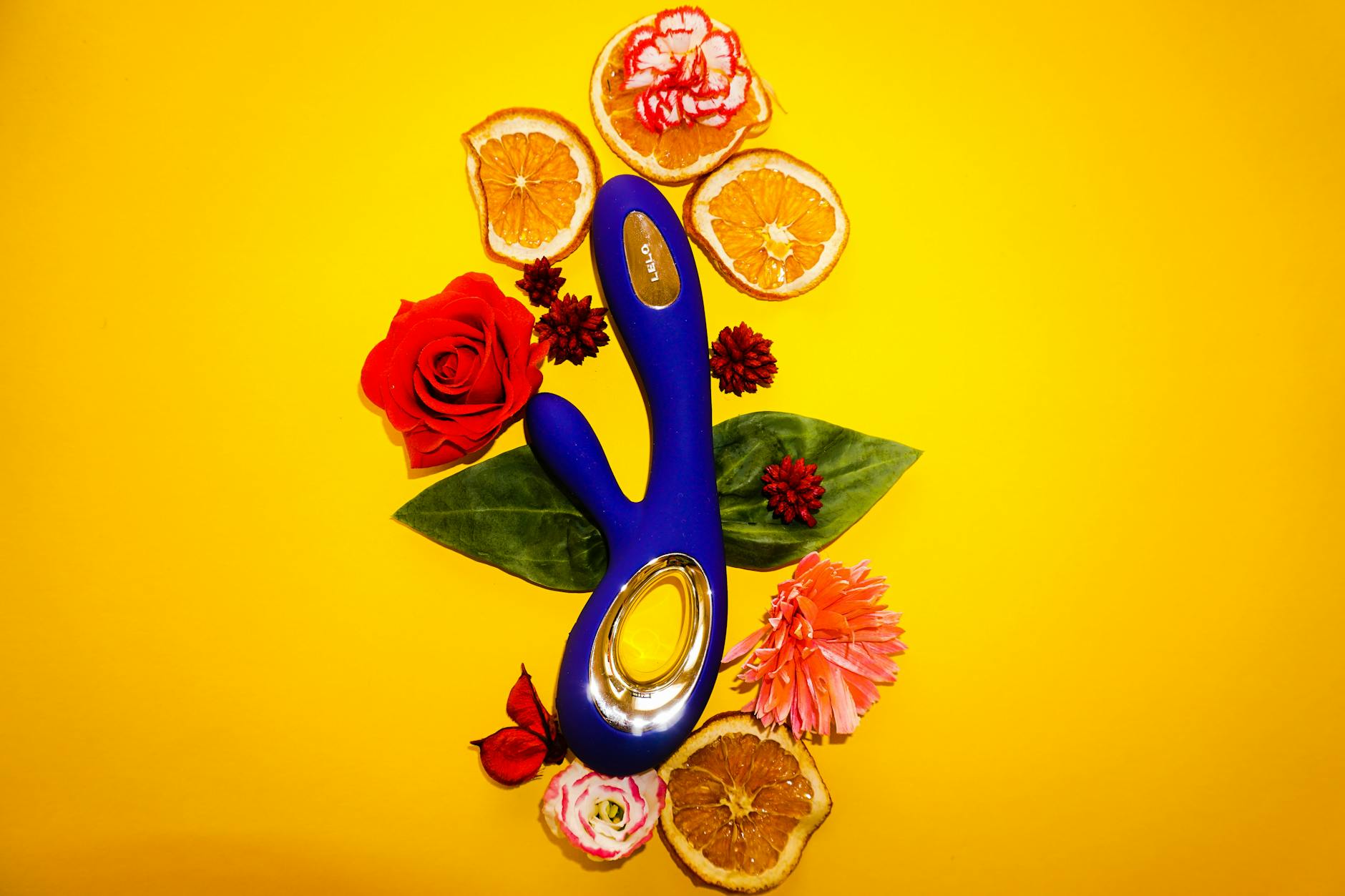 A yellow lemon clitoral vibrator surrounded by fresh flowers and citrus on a bright yellow background